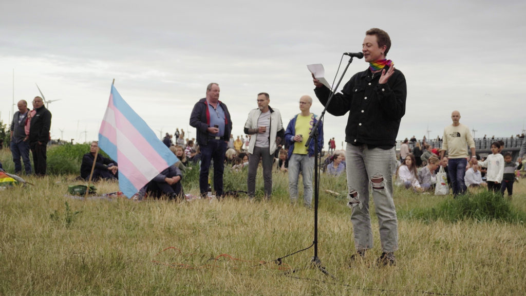 Rei McKay Mansa holdet tale på en græsplæne ved siden af et Trans Pride flag.