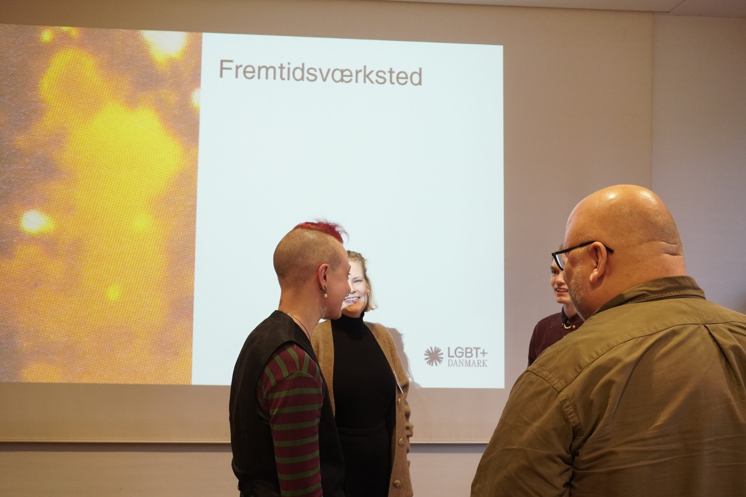 Deltagere til LGBT+ Danmarks årskonference taler sammen foran et slideshow med teksten "Fremtidsworkshop" på
