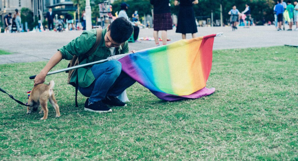Ung person på græsplæne med regnbueflag, klapper en lille hund.