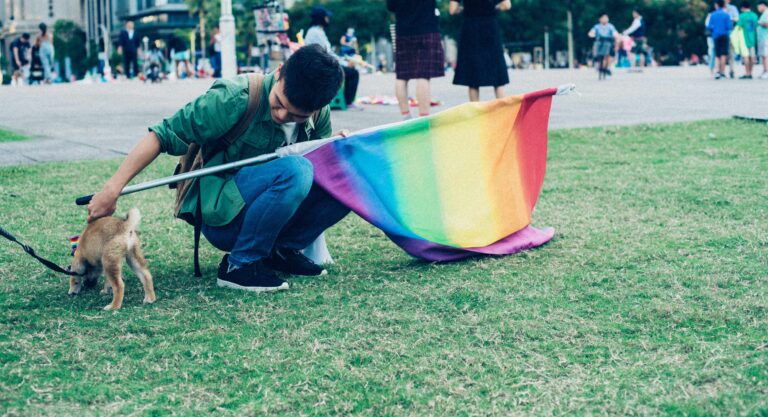 Ung person på græsplæne med regnbueflag, klapper en lille hund.
