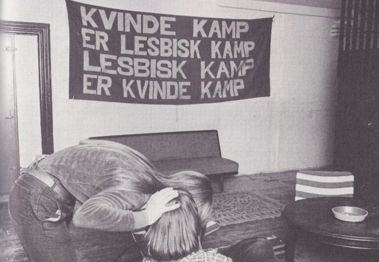 Billede fra 70'erne fra en stue med to mennesker, der sidder under et banner, hvorpå der står et lesbisk feministisk slogan "kvindekamp er lesbisk kamp, lesbisk kamp er kvindekamp