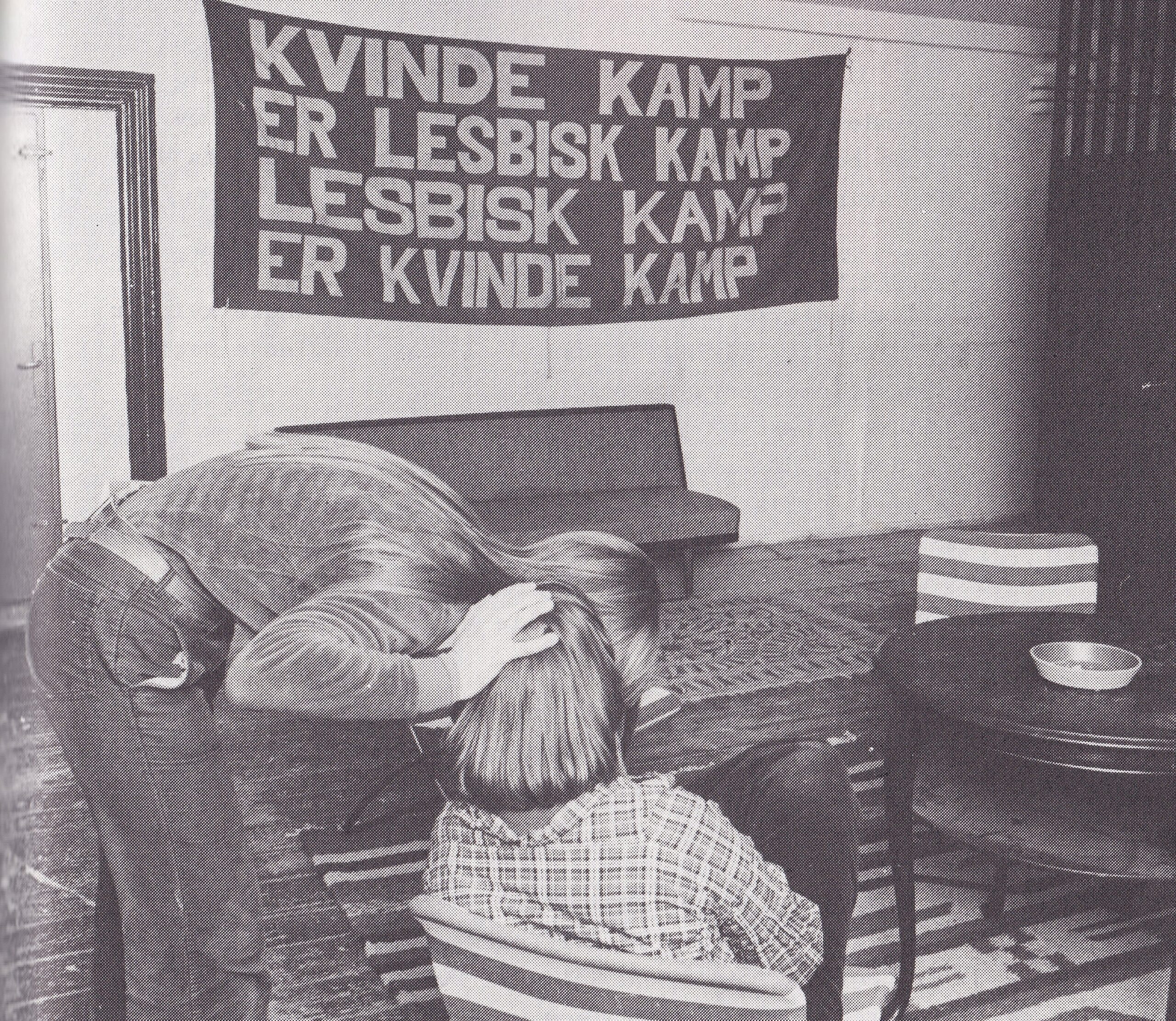 Billede fra 70'erne fra en stue med to mennesker, der sidder under et banner, hvorpå der står et lesbisk feministisk slogan "kvindekamp er lesbisk kamp, lesbisk kamp er kvindekamp
