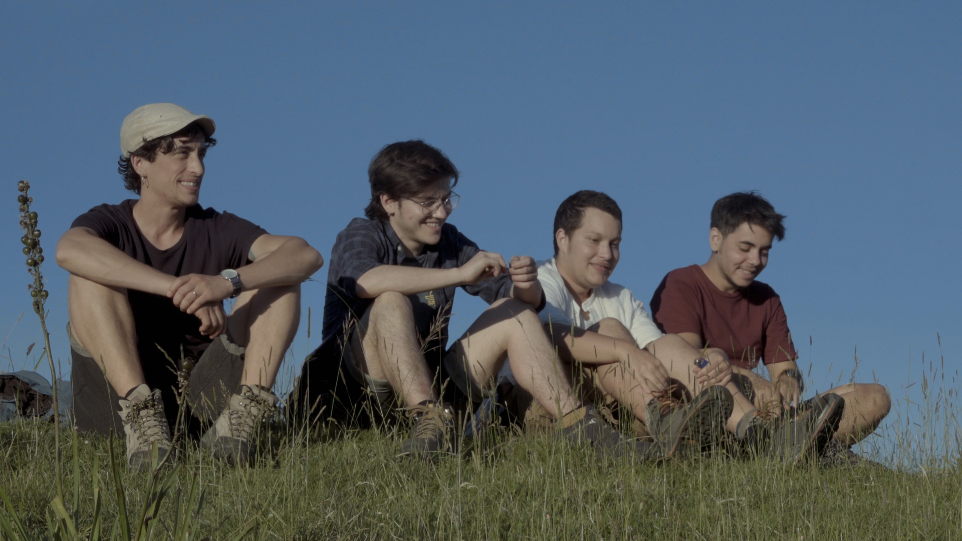 Still fra filmen Into My Name af fire transmænd der sidder i solen på en græstop med en blå himmel bag sig.